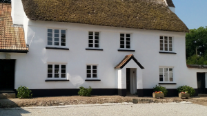 White traditional thatched cottage with black trim and multiple windows surrounded by greenery.