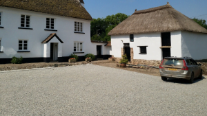 Two traditional thatched roof buildings with a gravel courtyard and a parked car in front.