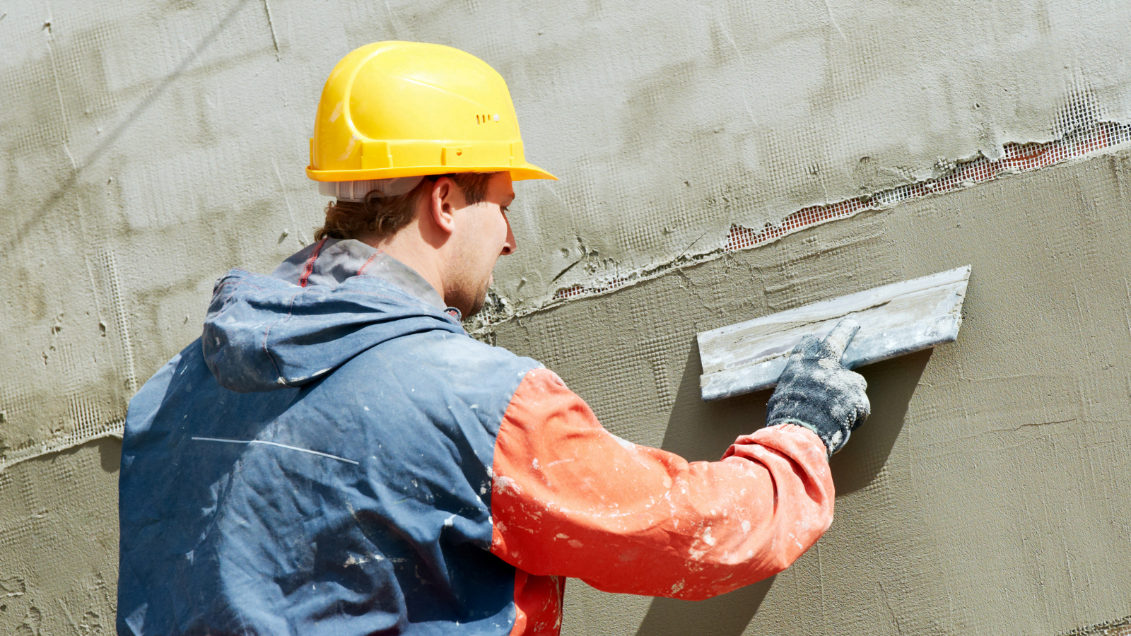 Construction worker in a yellow hard hat applying render to a wall with a trowel.