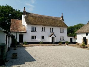 A traditional thatched cottage with white walls and a gravel courtyard, surrounded by greenery.
